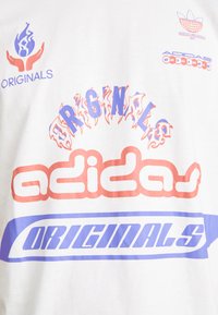 Balta medvilninė marškinėlių su spalvingais grafikos elementais, įskaitant raudoną ir mėlyną tekstą "adidas", "ORIGINALS" ir ugnies raštus.