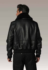 Homme portant une veste bomber en cuir noir avec un col en shearling, vu de dos sur un fond gris uni.