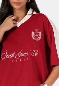 Rode trui met een witte kraag, voorzien van een logo op de linkerborst en "Sixth June, Co. PARIS" in het wit geborduurd. Soepele textuur.