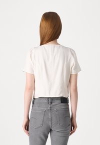 Top corto a maniche corte in cotone crema con una texture liscia. Presenta un colletto rotondo e dettagli di cucitura sottili. Abbinato a jeans grigi.