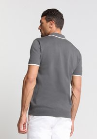Grijze polo met korte mouwen, witte kraag en mouwafwerking, gebreide textuur en uitsneden aan de zijkanten, gedragen met witte shorts.