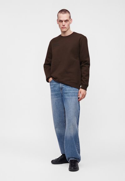 Homme aux cheveux courts, portant un sweat-shirt marron foncé, un jean bleu clair ample et des baskets noires, debout devant un fond clair uni.