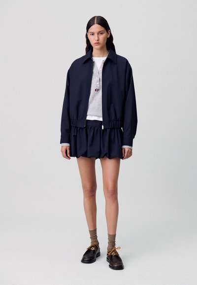 Blouson aviateur bleu marine avec poignets élastiques, associé à un haut gris à col rond et un short bleu marine à volants, complété par des chaussures foncées et des chaussettes marron.