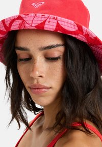 Červený klobúk s širokým okrajom, ktorý je pokrytý vzorom ružových listov. Vyrobený z jemnej bavlny. Malé srdiečkové logo vyšívané na prednej strane.