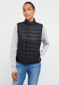 Czarna puchowa kamizelka z wysokim kołnierzem, pikowanym wzorem i subtelnym logo. Noszona na jasnoszarej koszulce z długim rękawem. Luźny fason z niebieskimi dżinsami.