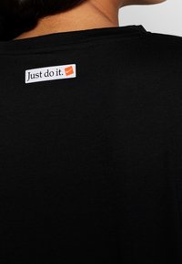 Czarna koszulka z gładką fakturą, z białą metką na plecach, na której widnieje napis "Just do it." oraz małe pomarańczowe logo Nike.