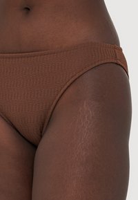 Brown bikini bottom in textured fabric, featuring a low-rise design with a smooth waistband, showcasing subtle stitching details.
