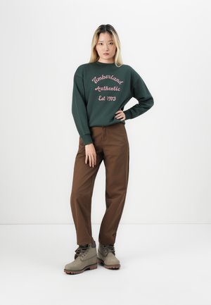 Grønn hettegenser med teksten "Timberland Authentic Est 1973", brune bukser, og grå støvler med svarte snørebånd, vist mot en hvit bakgrunn.