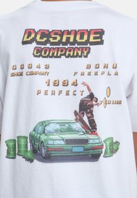 Bílý bavlněný tričko s grafikou zeleného auta, skateboardisty a herního textu v vícebarevném pixelovaném písmu.