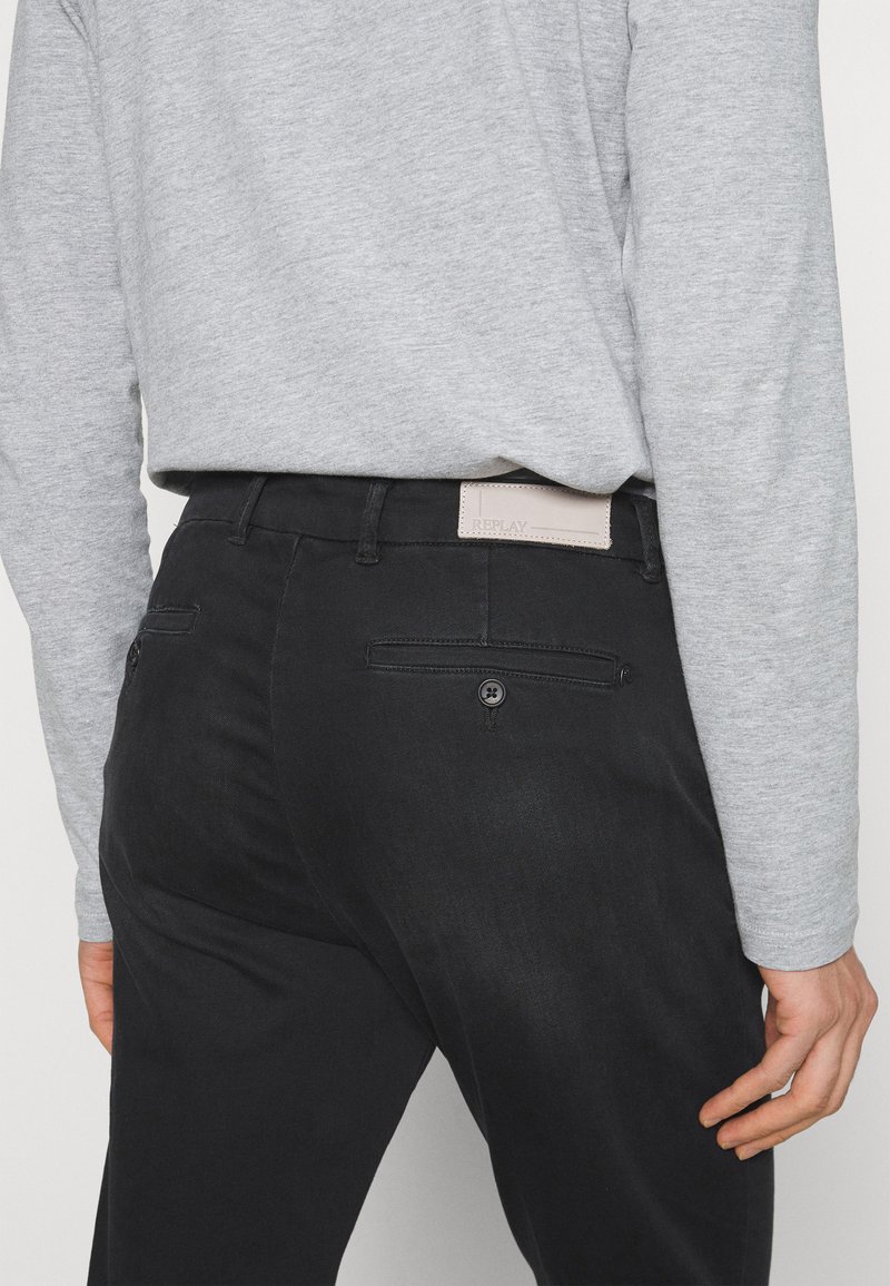 Schwarze Hose mit glatter Textur, ausgestattet mit Gesäßtaschen und einem beigen Lederetikett. Tragen mit einem grauen Langarmhemd.