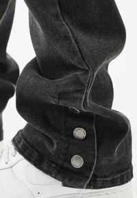 Zwarte denim jeans hebben een relaxed fit, met een omslagdetail en metalen knopen. Ze worden gecombineerd met witte sneakers.