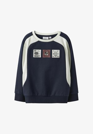 Tumši zila džemperis ar krēmkrāsas akcentiem, kurā ir trīs izšūti plāksteri ar hokeja elementiem. Apaļš kakla izgriezums un ribota apakšdaļa.