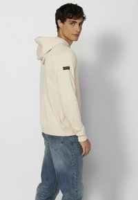 Haut à capuche en tricot beige avec une finition texturée, doté d'une capuche à cordon de serrage et d'un panneau en maille sur le côté, avec un écusson logo sur la manche.