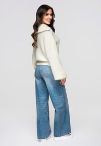 Pull en maille beige clair avec un grand col, associé à un jean bleu à jambes larges. La tenue présente une coupe décontractée et des manches retroussées.