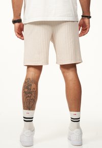Beige strukturierte Shorts mit vertikalem Rippenmuster, getragen mit weißen Socken mit schwarzen Streifen und weißen Turnschuhen. Sichtbares Tattoo am Bein.