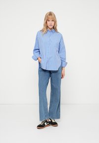 Világoskék gombos ing sötétkék függőleges csíkokkal, széles szárú farmernadrággal és fekete sportcipővel, barna talppal.