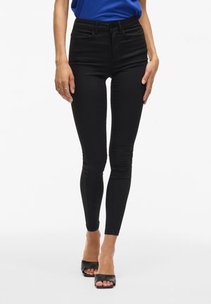 Femme portant un jean skinny noir taille haute, un haut bleu et des talons ouverts noirs, debout devant un fond blanc.