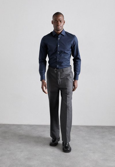 Chemise bleu marine à boutons associée à un pantalon gris ; présente une texture lisse, une coupe ajustée et un léger éclat ; complétée par des chaussures noires.