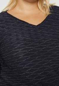 Mörkblå topp med V-ringning, prydd med ett strukturerat vågmönster som accentueras med metalltrådar för en subtil glimrande effekt.