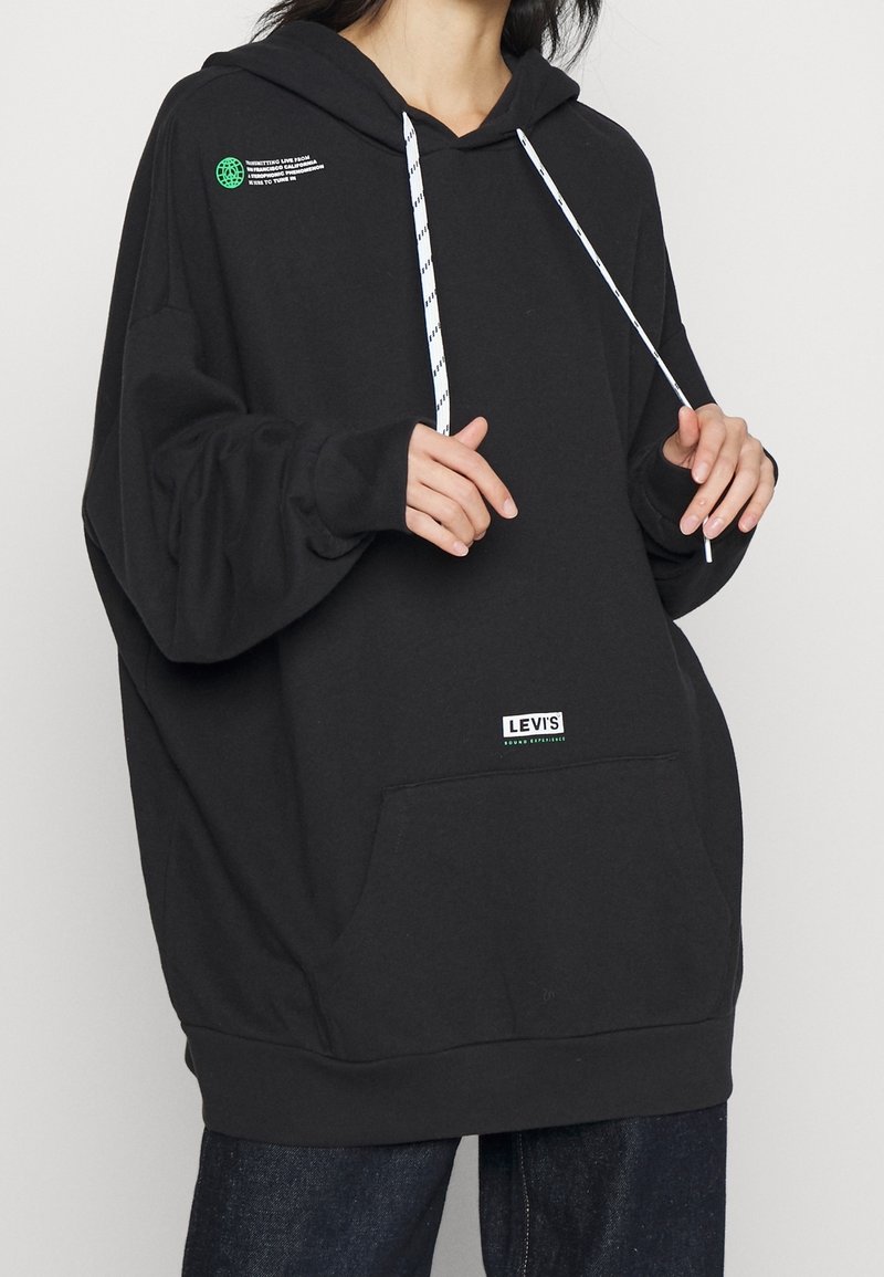Femme portant un sweat à capuche noir oversize Levi’s avec des cordons blancs et une poche avant, debout devant un fond clair uni.