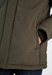 Olivengrøn pufferjakke med en blød, struktureret overflade, der har forlommer, en høj krave og et rent, enkelt design.