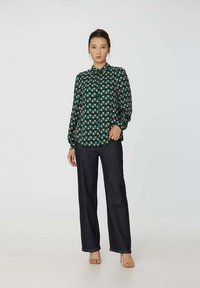 Blouse verte avec un motif géométrique de carrés roses et blancs, design à boutons, et manches longues bouffantes. Associée à un pantalon en denim foncé.