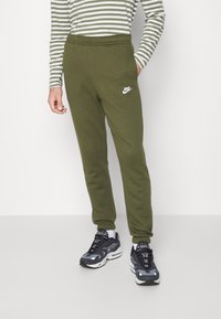 Olivgröna Nike mjukisbyxor med elastisk midja och muddar, med en vit Nike-logga på övre låret. Matchas med svarta sneakers.