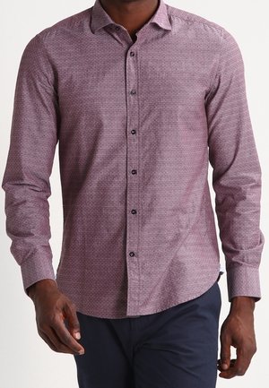 Mężczyzna noszący dopasowaną bordową koszulę na guziki z długim rękawem w wzory oraz ciemnoniebieskie spodnie, pokazany od szyi do połowy uda.