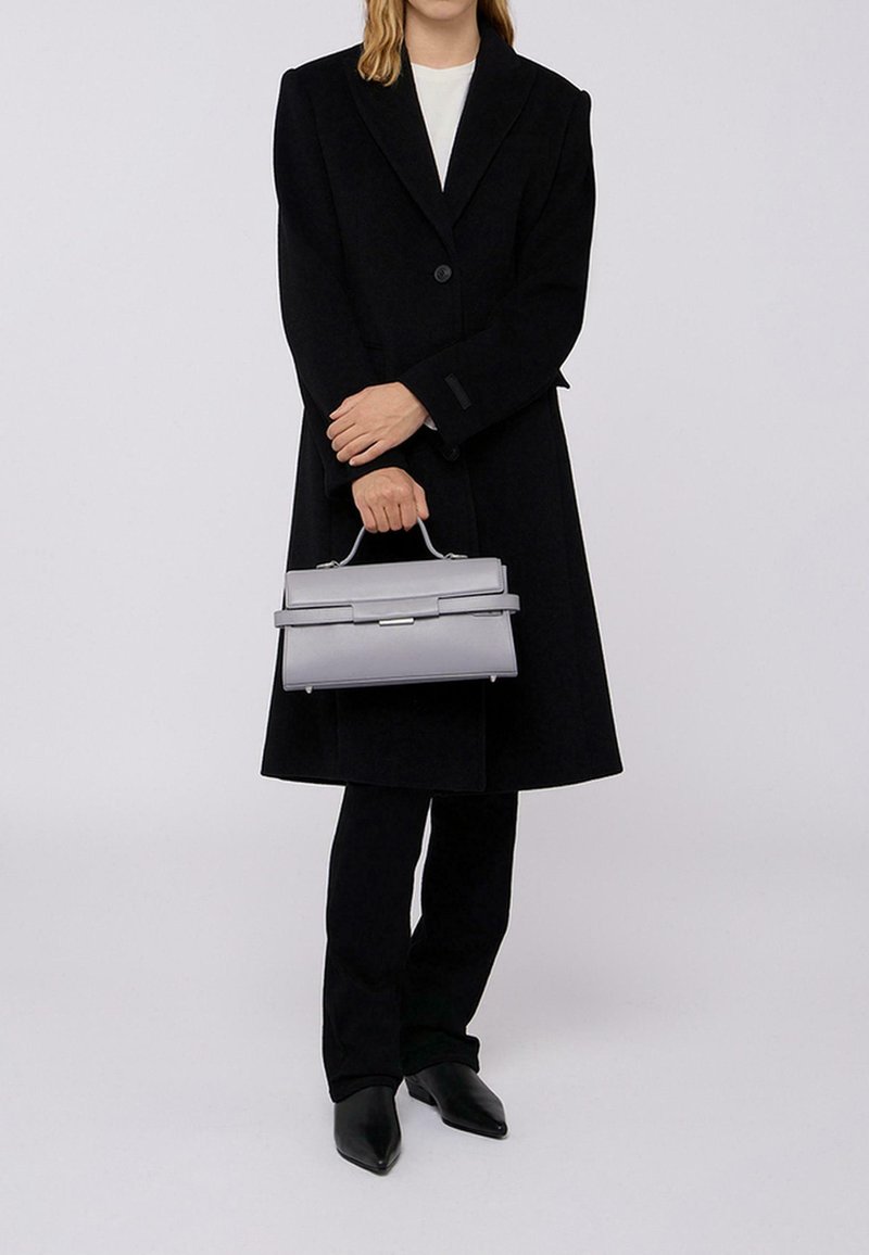 Borsa strutturata grigia con manico superiore, caratterizzata da un design elegante, una texture liscia e una sottile tracolla sul davanti. Indossato un cappotto nero e pantaloni.