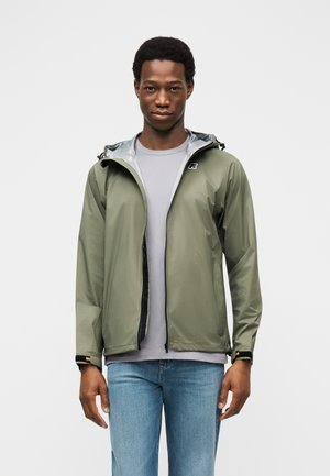 Mladý muž oblečený v olivovo zelenej nepremokavej bunde cez sivé tričko a svetlomodré rifle, stojaci na jednoduchom bielom pozadí.