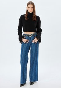 Schwarzer gestrickter Crop-Pullover mit fransigen Ärmeln, kombiniert mit hochtaillierten blauen weit geschnittenen Jeans, die ein dezentes Muster und einen schmalen Gürtel aufweisen.