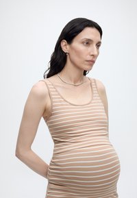 Έγκυος γυναίκα που φοράει αμάνικο φόρεμα με μπεζ και λευκές ρίγες, στέκεται με τα χέρια πίσω από την πλάτη και κοιτάζει μπροστά με ουδέτερη έκφραση.