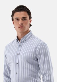 Hellblaues Button-Up-Hemd mit vertikalen weißen Streifen, langen Ärmeln, einer Brusttasche und silbernen Knöpfen an einem strukturierten Kragen.