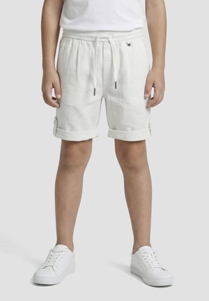 Niño con pantalones cortos blancos con cordón y bajos remangados, y zapatillas blancas, de pie frente a un fondo claro y liso.