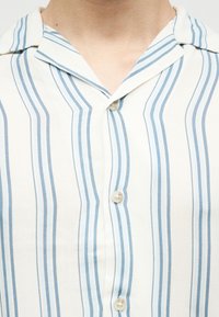 Κοντινό πλάνο ενός ατόμου που φοράει λευκό πουκάμισο με κάθετες ανοιχτό μπλε ρίγες και διπλωμένο γιακά, κουμπωμένο μπροστά.