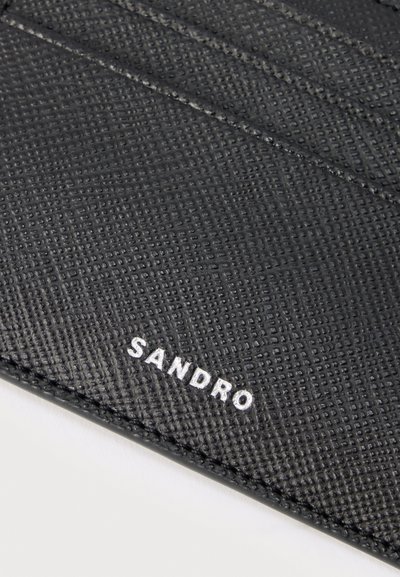 Κοντινό πλάνο υφασμένου μαύρου δερμάτινου πορτοφολιού με την επωνυμία "SANDRO" τυπωμένη με λευκά γράμματα κοντά στην κάτω άκρη.