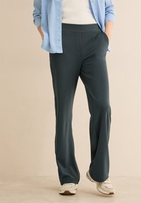 Ciemnozielone, lekkie spodnie o gładkiej teksturze, z elastycznym paskiem w talii, przednimi kieszeniami i luźnym krojem, połączone z białymi butami sportowymi.