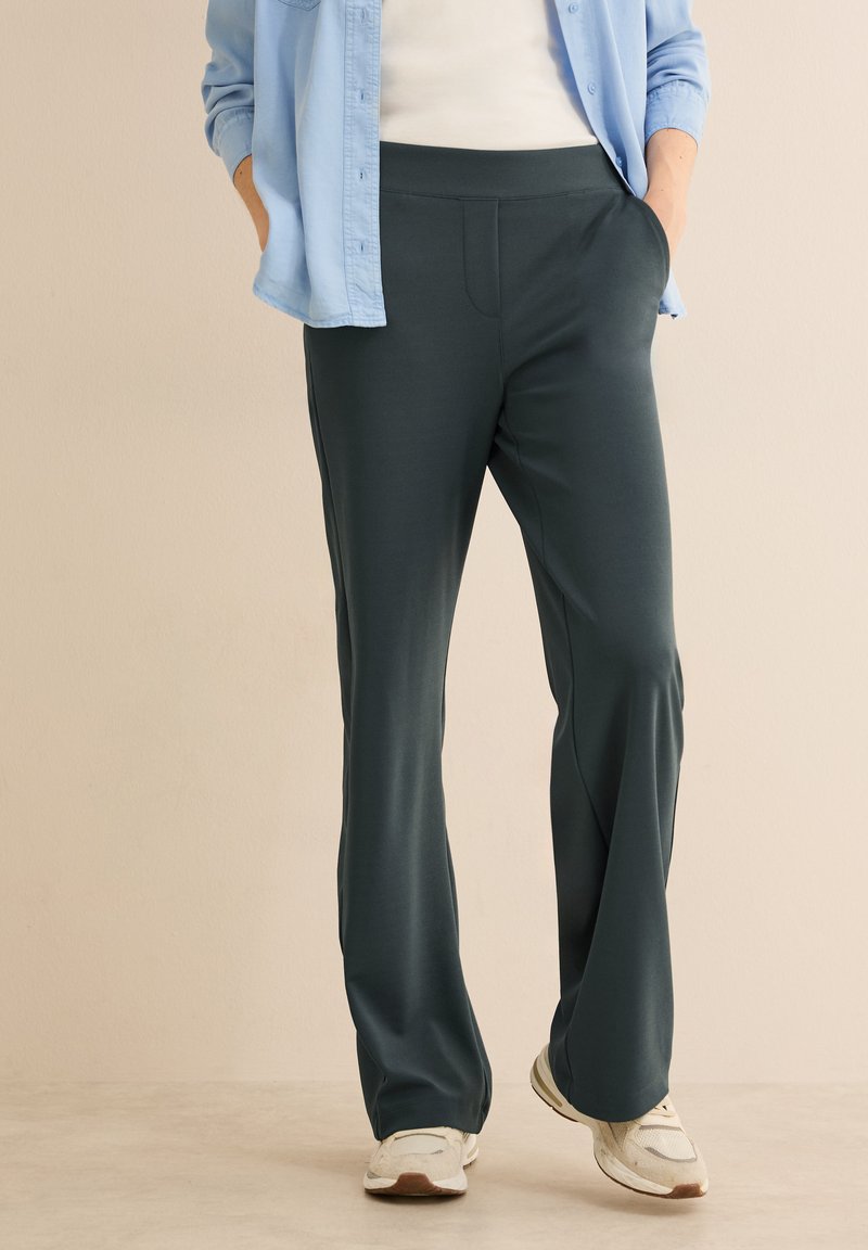 Ciemnozielone, lekkie spodnie o gładkiej teksturze, z elastycznym paskiem w talii, przednimi kieszeniami i luźnym krojem, połączone z białymi butami sportowymi.