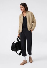 Beiger Sweatshirt mit Reißverschluss, schwarzes T-Shirt, lockere schwarze Hose, beige Sneakers, trägt eine schwarze Sporttasche; lässig, entspannter Schnitt.