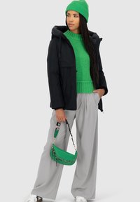 Giubbotto a vento nero, maglione lavorato a maglia verde, pantaloni larghi grigi e una borsa verde con una tracolla a fantasia. Modella che indossa un berretto verde.