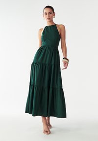 Zelena maksi obleka iz lahkega materiala, ki vključuje halter ovratnik, oprijet zgornji del in obiščeno krilo. Preprost dizajn brez vzorcev.