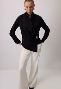 Melnā blūze ar gludu tekstūru un zelta akcentu jostasvietā, apvienota ar baltām platām biksēm.