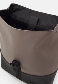 Sac gris et noir en matériau synthétique lisse, doté d'une ouverture supérieure, d'une poche intérieure et d'un accent de sangle noire.