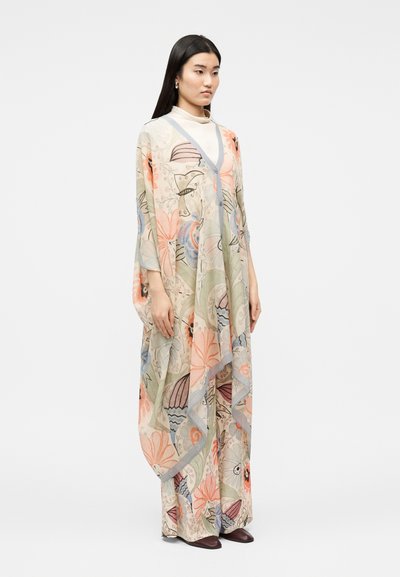 Γυναίκα που φοράει ένα ρέον χιτώνα με λουλουδάτο μοτίβο σε παστέλ αποχρώσεις, πάνω από παντελόνια με φαρδιά μπατζάκια σε ταιριαστό χρώμα και σκοτεινά παπούτσια, στέκεται μπροστά σε ένα απλό φόντο.