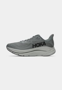 Sapatilha de corrida atlética cinza com sola branca espessa e logo preto "HOKA" na lateral, apresentada contra um fundo branco liso.