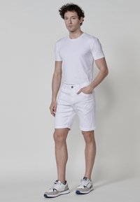 Camiseta de algodón blanca y pantalones cortos de mezclilla blanca con detalles desgastados; modelo que lleva zapatillas blancas con acentos en gris y navy; fondo neutro.