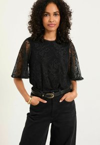 Schwarzes Spitzenoberteil mit kurzen Ärmeln und gewelltem Saum, getragen mit hochtaillierten schwarzen Hosen und einem schwarzen Gürtel mit einer goldenen Schnalle.