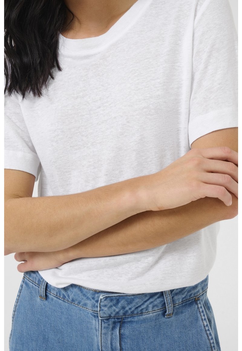 Personne aux longs cheveux foncés portant un t-shirt blanc uni à manches courtes et un jean bleu, bras croisés à la taille.