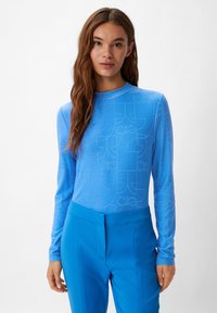 Maglietta a maniche lunghe di colore azzurro chiaro con un motivo sottile che presenta forme arrotondate. Il materiale appare liscio e aderente. Abbinata a pantaloni blu coordinati.