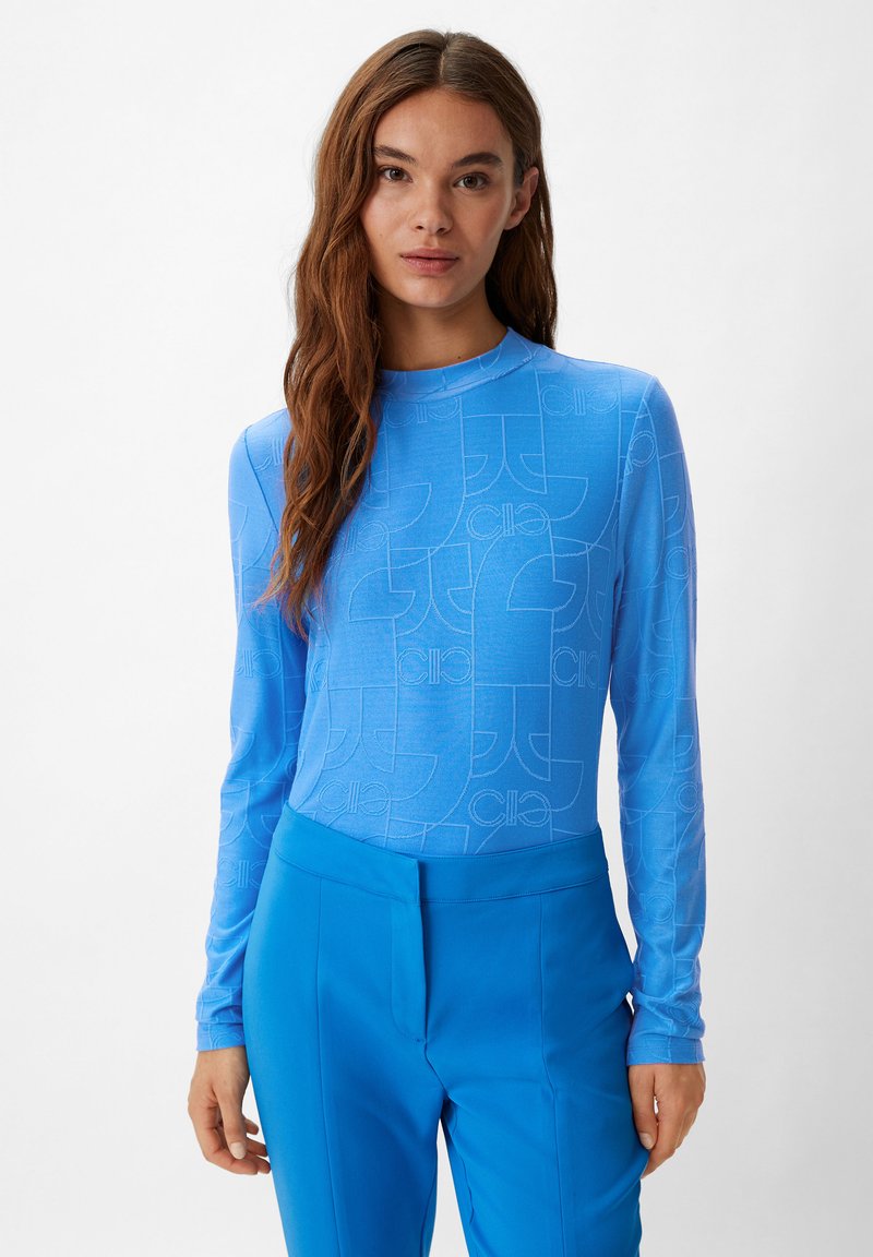 Maglietta a maniche lunghe di colore azzurro chiaro con un motivo sottile che presenta forme arrotondate. Il materiale appare liscio e aderente. Abbinata a pantaloni blu coordinati.